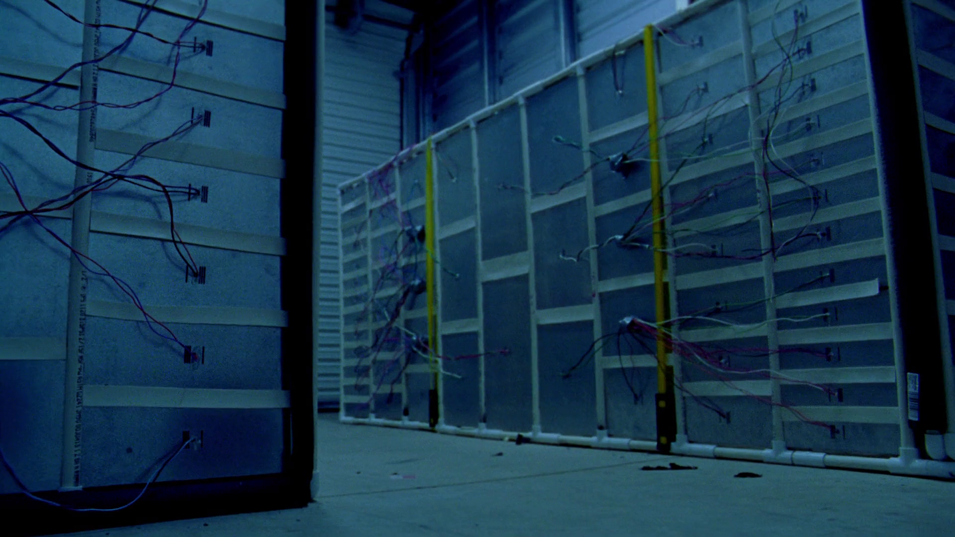 A stark, clinical shot of a grey, unadorned metal apparatus, roughly the size of a refrigerator, sitting in the center of a mundane storage unit.