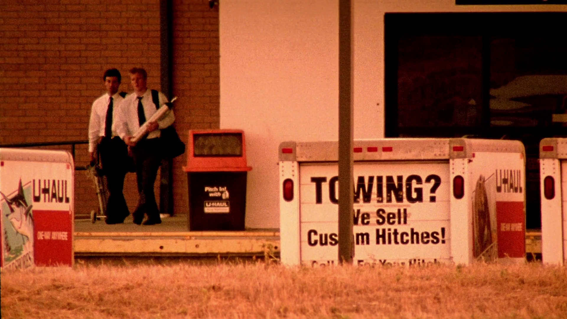 A point-of-view shot from a distance, looking across a field as Aaron watches his exact double exit the storage facility with Abe.