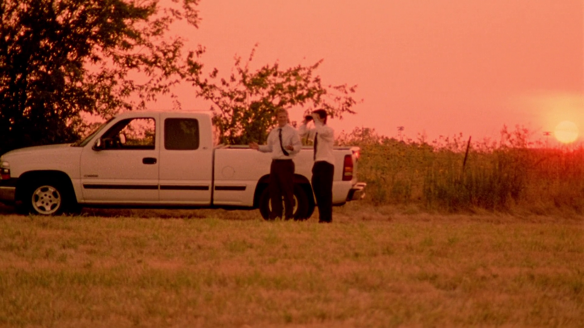 Two men, Aaron and Abe, stand beside a white pickup truck in a grassy field under a vivid reddish-orange sunset sky. One of the men holds binoculars to his eyes, looking intently across the field to the right, observing their doubles in the distance.