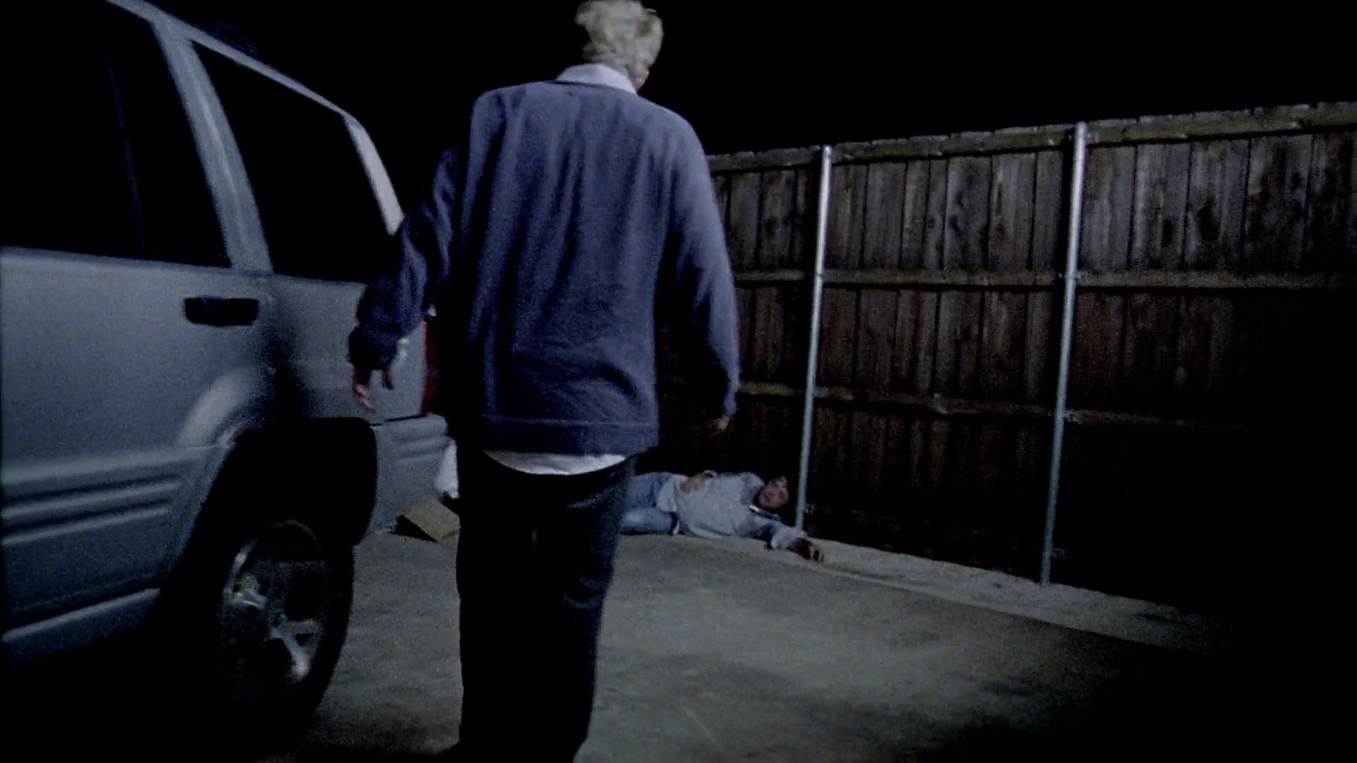 A dark, nighttime shot showing a man lying collapsed on a concrete driveway between a silver SUV and a wooden privacy fence. Abe, seen from behind in the foreground, walks toward the unconscious figure, identified as Granger.
