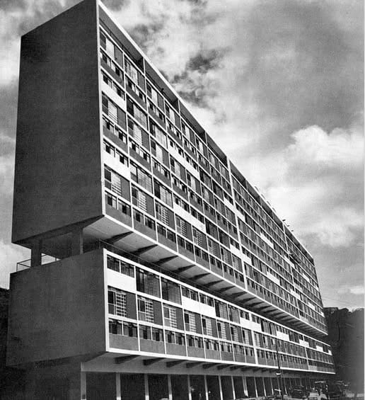 A low-angle black-and-white photograph from the mid-1950s showing a massive modernist apartment block in Caracas, Venezuela. The building features a rigid geometric facade with recessed balconies and sun-shades, raised on concrete pilotis (pillars) at the ground level, looming over the viewer like a "giant sideways domino" against a cloudy sky.