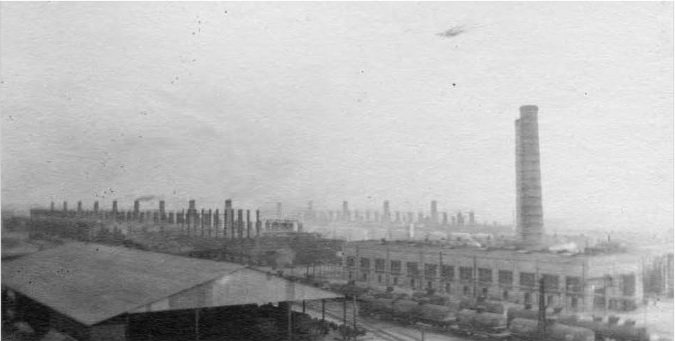 A panoramic, grainy black-and-white photograph of a vast oil refinery complex. Dozens of tall smokestacks line the horizon above long factory buildings, with a row of rail tank cars visible on tracks in the foreground.