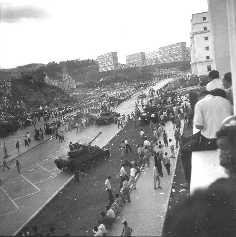 A high-angle black-and-white photograph of the January 1958 coup in Caracas. Military tanks move down a wide avenue lined with the "modernistic" superblocks, while civilians watch from the foreground ledges, witnessing the moment the "dictatorship’s housing project" became a symbol of revolution.