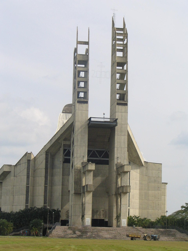 A color photograph of the massive National Basilica of Our Lady of Coromoto in Venezuela. The Brutalist structure rises sharply from the flat landscape, composed of soaring, angular grey concrete walls that converge like the prow of a ship or a modern pyramid against a bright blue sky.