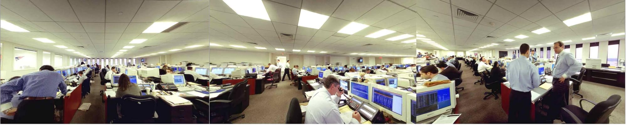 A wide, panoramic interior shot of a large, busy office space filled with rows of computer monitors and desks. Numerous employees in business attire are seen working at their stations or standing and talking.