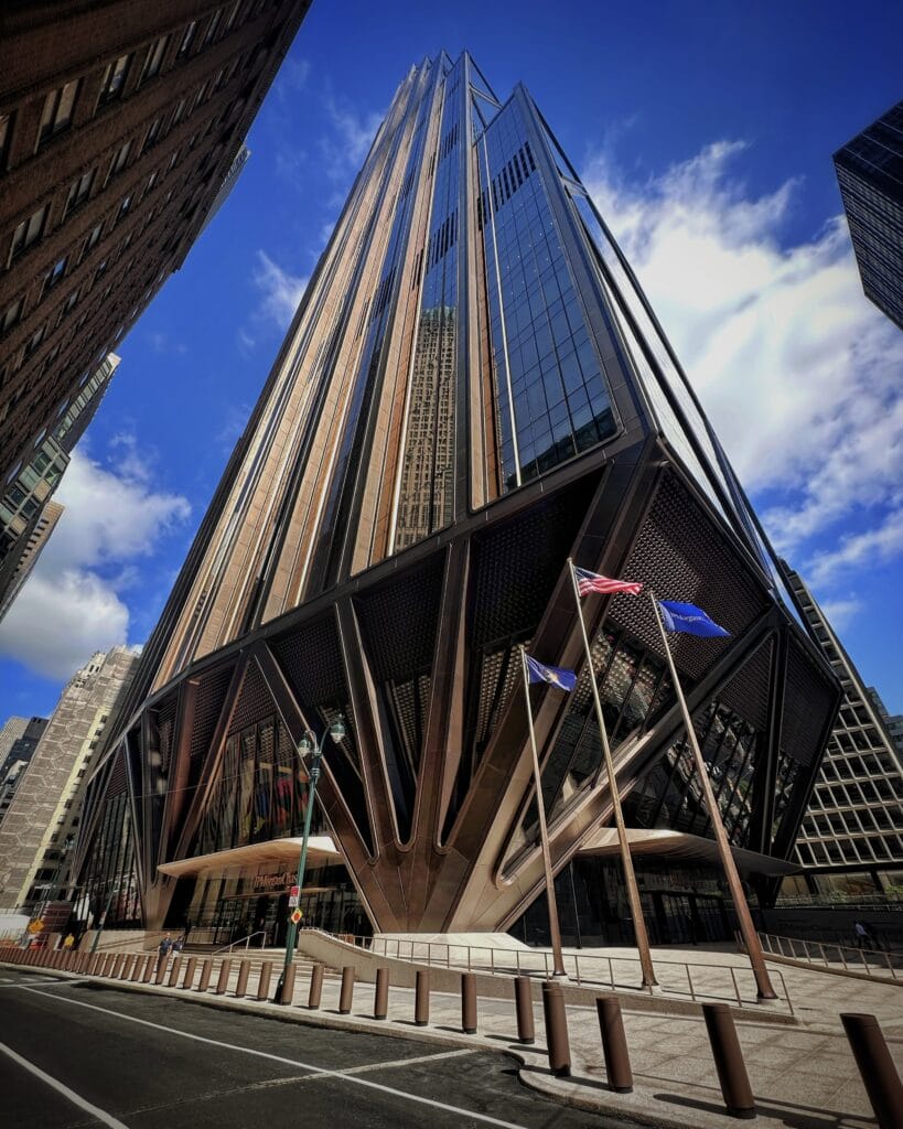 A dramatic, upward-looking view of the modern skyscraper JPMorgan Chase headquarters with a unique fan-like or V-shaped structural base. American and New York State flags fly in front of the entrance under a clear blue sky with light clouds.