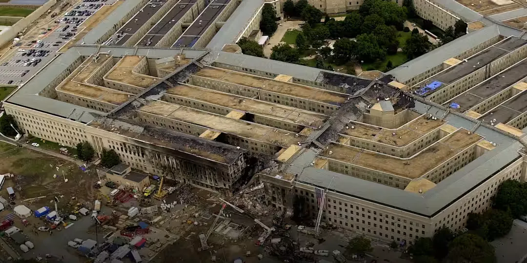 An overhead photograph showing a collapsed and scorched section of the Pentagon building. Debris and emergency vehicles are visible on the lawn surrounding the impact site where several rings of the structure were breached.