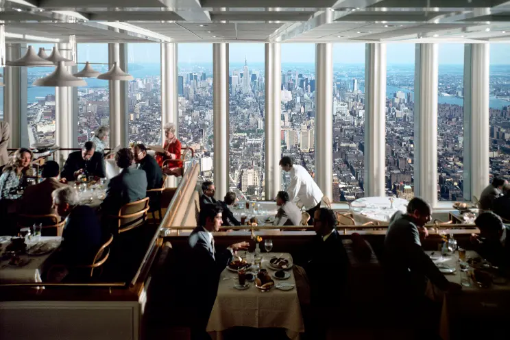 An interior photograph of Windows on the World, a high-end restaurant with floor-to-ceiling windows. People are seated at tables set with white linens, enjoying a meal while looking out at a sprawling panoramic view of New York City from top of the North Tower.