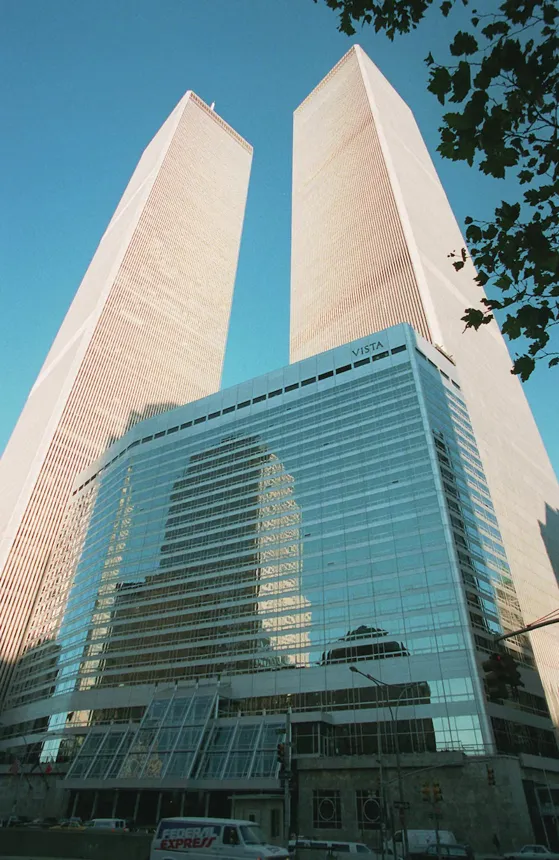 A daytime, upward-looking shot of the North and South Towers of the original World Trade Center against a clear blue sky. A shorter, modern glass building labeled "VISTA" sits at the base, and an "EXPRESS" delivery van is visible at street level.