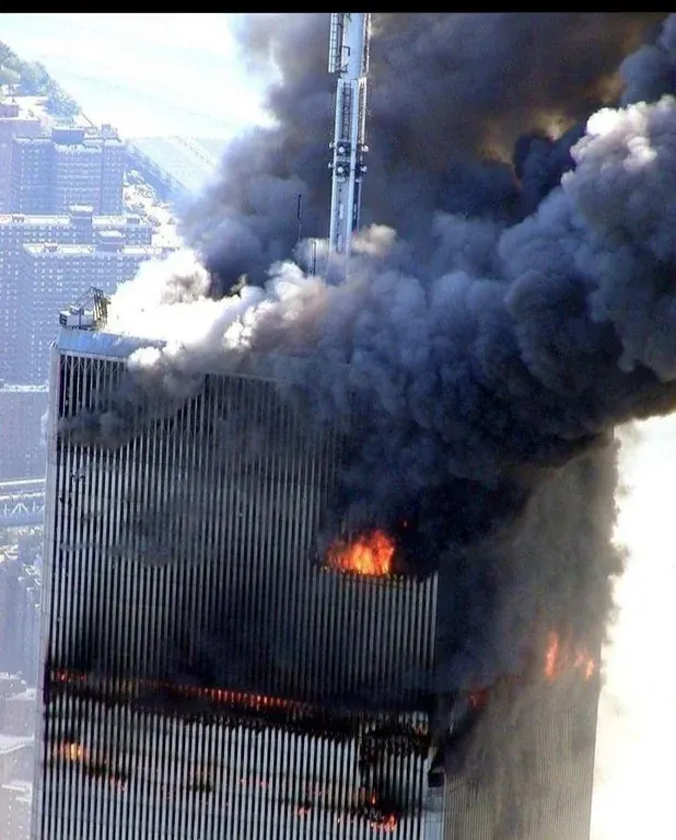 A photograph of the upper floors of a World Trade Center tower. Thick black smoke billows from a large, gaping hole in the side of the building where fire is visible within the structure.