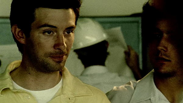 A wide shot of a vast warehouse where Aaron stands directing a group of workers constructing a colossal metal frame.